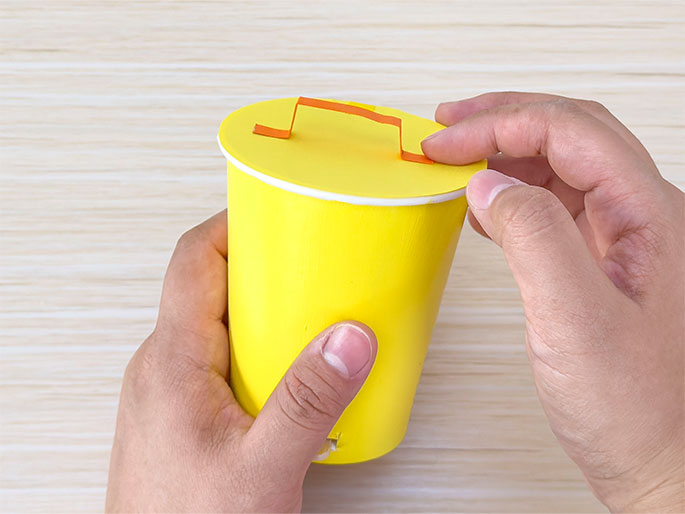 Two hands hold a bright yellow paper cup with a yellow circular lid over a light woodgrain background. An orange paper handle, shaped like a squared-off U, is attached to the lid. One hand holds the cup while the other adjusts the lid and handle. The image shows a completed or nearly completed craft project resembling a small bucket or container made from a paper cup.