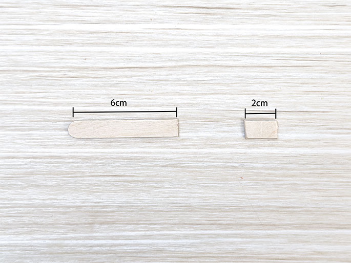 Two small pieces of light-colored wood or craft stick material lie on a light woodgrain surface. The larger piece on the left is 6cm long and has rounded ends. The smaller piece on the right is 2cm long and appears to be a section cut from a similar stick, with straight cut ends. Lines and labels indicate the measurements of each piece. The image illustrates the preparation of specific-sized wooden pieces for a craft project.