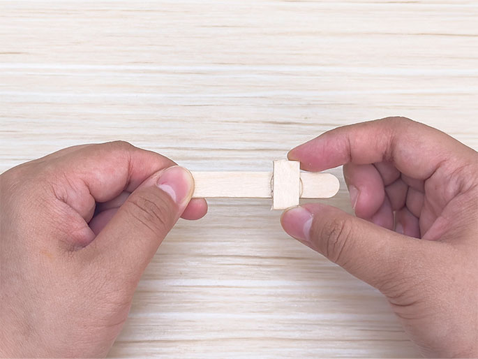 Two hands hold two small pieces of light wood, likely craft sticks or popsicle stick pieces, against a light woodgrain background. A smaller piece is attached perpendicularly to a longer piece, forming a T shape. It appears the smaller piece has been glued or otherwise fastened to the longer one. The hands are carefully holding the pieces together, likely ensuring the bond sets. The image shows the process of joining two small wooden pieces for a craft project.