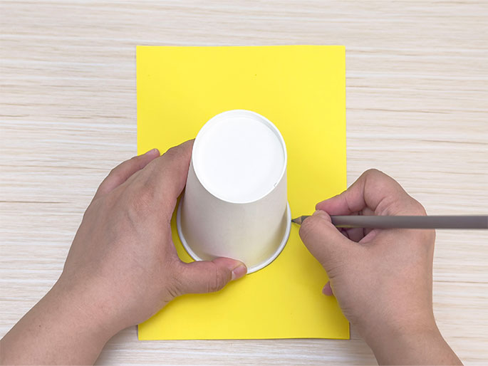 Two hands are shown tracing the bottom of a white paper cup onto a piece of bright yellow construction paper. The cup is placed upside down on the paper, and a pencil is being used to carefully trace around the rim. The image captures the process of creating a circle template using a cup and pencil on colored paper, likely for a craft activity.