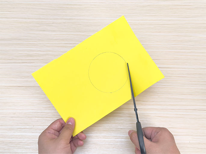 A pair of hands is cutting out a circle from a piece of bright yellow construction paper with scissors. A pencil-drawn circle is visible on the paper, guiding the cutting process. The hands hold the paper and scissors against a light woodgrain background. The image shows the step of cutting a pre-drawn circle from construction paper, a common task in craft projects.