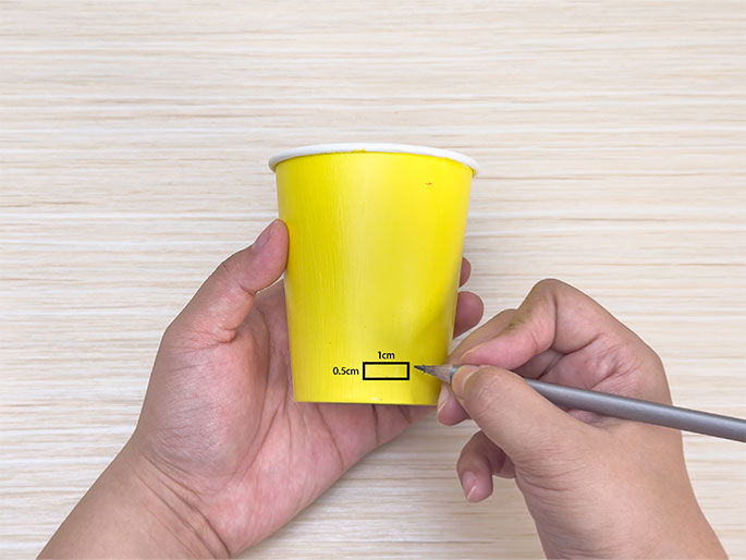 Two hands hold a bright yellow paper cup against a light woodgrain background. One hand holds the cup steady while the other hand uses a pencil to draw a small rectangle near the bottom edge of the cup. The rectangle is labeled with dimensions: 1cm long and 0.5cm wide. The image demonstrates marking a specific area for cutting or modification on a paper cup as part of a craft project.