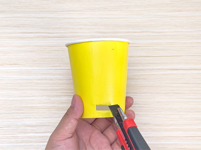A hand holds a bright yellow paper cup against a light woodgrain background. A utility knife with a red and black handle is being used to carefully cut a rectangular slot in the side of the cup. The image demonstrates the process of modifying a paper cup by cutting a precise opening, likely for a craft or DIY project.