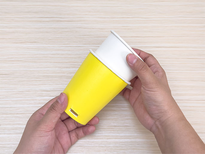 Two hands hold a bright yellow paper cup with a rectangular slot cut into its side and a plain white paper cup against a light woodgrain background. The white cup is positioned slightly inside and on top of the yellow cup, as if being placed over it. The image shows two paper cups being used in a craft project, likely being combined or modified in some way.