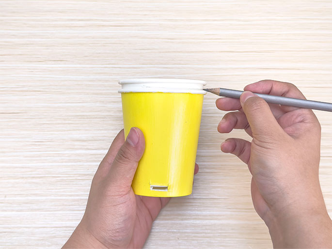 Two hands hold a bright yellow paper cup with a rectangular slot cut into its side and a white paper cup rim against a light woodgrain background. The white rim is placed on top of the yellow cup, and a pencil is marking a line around the inside edge of the white rim. The image demonstrates the process of marking a cutting line on a paper cup using a second cup's rim as a template, likely for a craft activity.
