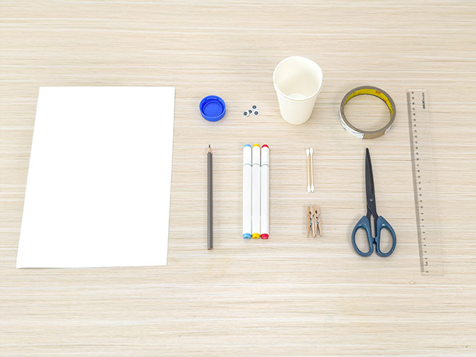 A flat lay arrangement of various craft supplies is displayed on a light wood grain table. The items include a blank sheet of white paper, a gray pencil, three white markers with colored caps, a small blue bottle cap, a pair of googly eyes, a small white paper cup, a roll of clear tape, a pair of small scissors, a wooden ruler, two cotton swabs, and a small wooden clothespin. These materials appear to be gathered for a children's craft or DIY project.