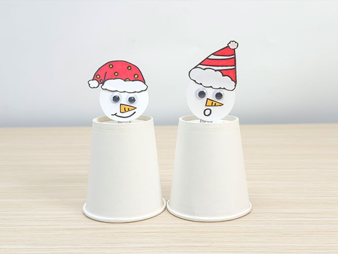 Two cheerful snowman figures, crafted from paper, are perched atop white paper cups on a light wood-grained surface. Each snowman has a round white face with googly eyes and a carrot nose. The snowman on the left sports a red Santa hat with yellow dots and a smile. The snowman on the right wears a red and white striped Santa hat and has a surprised expression with an o shaped mouth. The image showcases a simple, festive craft project.