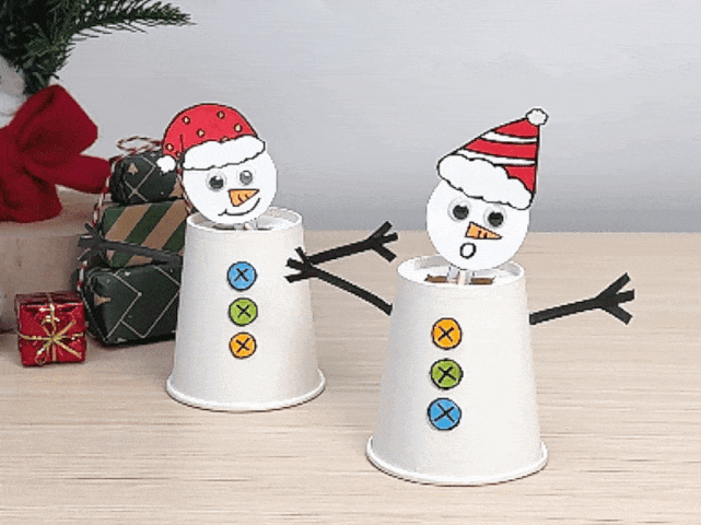 Two charming paper cup snowmen are displayed on a light wood-grained table with festive Christmas decorations in the background. The snowmen are made from white cups, each decorated with three colorful buttons marked with Xs. They have round white heads with googly eyes, orange carrot noses, and Santa hats. The snowman on the left wears a red Santa hat with yellow dots and has a smiling expression. The snowman on the right wears a red and white striped Santa hat and looks surprised. Both snowmen have outstretched arms made of black paper. Wrapped presents and a decorated Christmas tree are visible behind the snowmen, adding to the holiday scene.