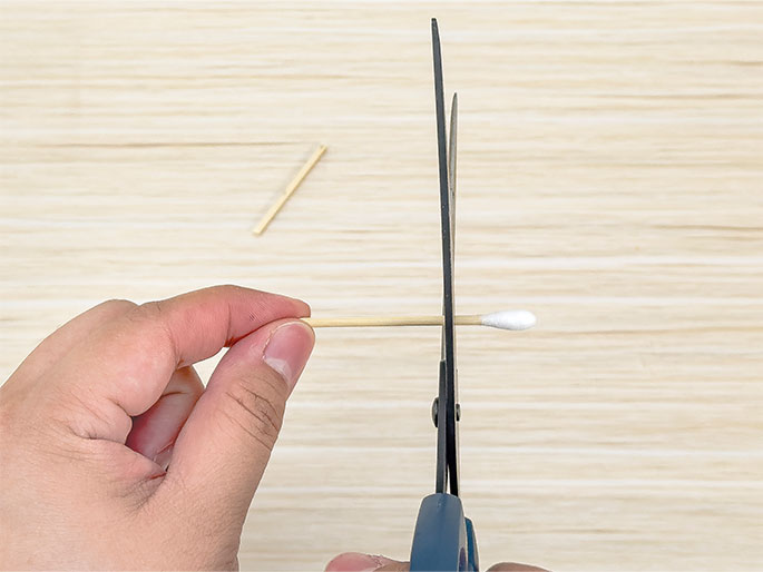 A hand holds a cotton swab horizontally positioned between the blades of a pair of open scissors, poised to cut the swab's wooden stick. The scissors have dark blue handles and metal blades. The background is a light wood-grained surface, and a small piece of a previously cut cotton swab stick lies on the surface to the upper left. The image demonstrates the action of cutting a cotton swab in half for a craft or other purpose.