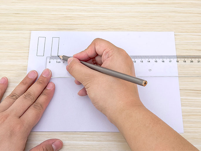 Two hands are shown interacting with a piece of white paper and a clear plastic ruler on a light wood-grained table. One hand holds a gray pencil and uses it to draw a straight line along the edge of the ruler. The other hand holds the ruler in place on the paper. Two rectangular shapes are lightly drawn in the upper left corner of the paper. The image suggests someone is following precise measurements for a drawing or craft project.