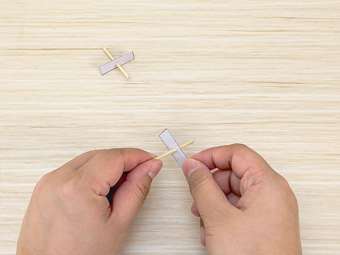 Two hands are assembling a small cross-shaped object made from thin wooden sticks and a small strip of clear tape on a light wood-grained surface. One completed cross lies on the table in the upper left corner, while the hands are in the process of taping the two sticks of the second cross together in the center of the image. This suggests a step-by-step illustration of a simple craft project.