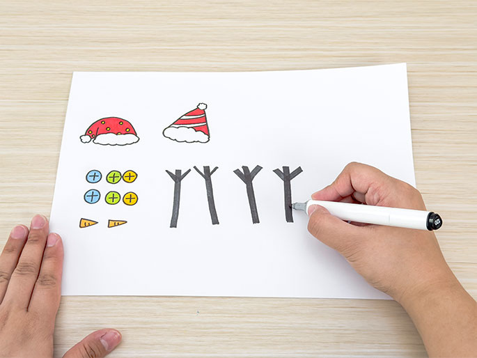 On a piece of white paper resting on a light wood-grained table, several simple drawings are depicted. These include two Santa hats (one red with white trim and one red with white stripes), two small orange carrot noses, six colorful buttons with plus sign details, and four dark gray stick-like shapes resembling bare tree branches. A hand holding a white marker is coloring in one of the tree branch shapes. The arrangement suggests the creation of a holiday-themed craft or drawing activity.