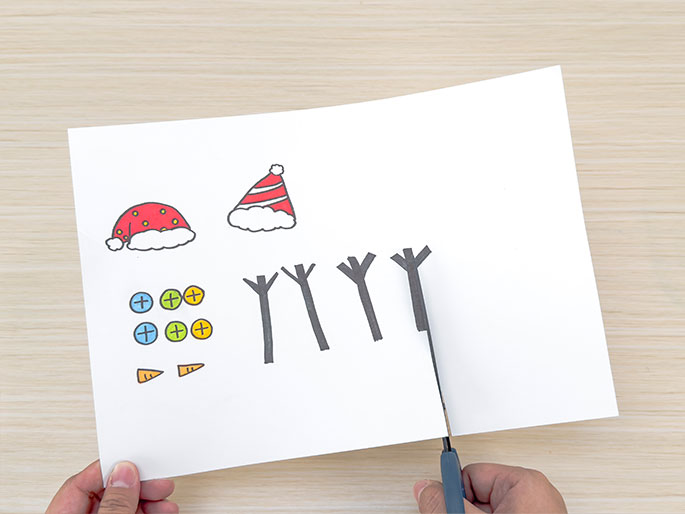 A pair of hands is cutting a piece of white paper with scissors. Drawn on the paper are various craft elements, including two Santa hats, colorful buttons, carrot noses, and four dark gray stick-like shapes, likely representing tree branches. The scissors, held in one hand, are in the process of cutting out one of the branch shapes. The other hand holds the paper steady. The image depicts a step in a crafting process, likely creating holiday-themed decorations.