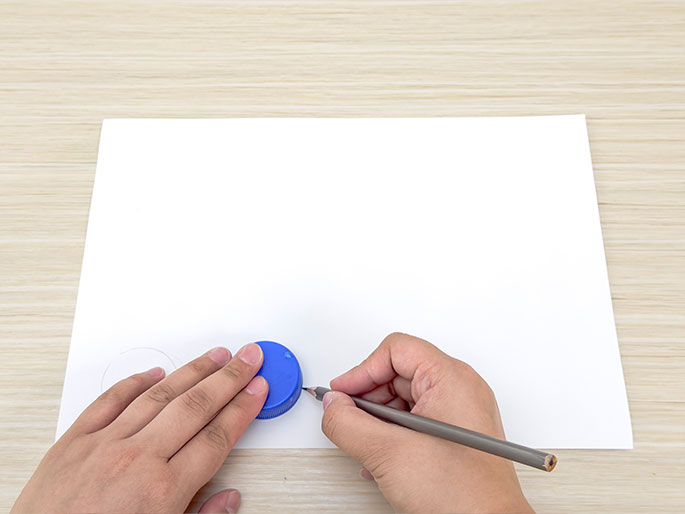 Two hands are shown tracing a circle on a piece of white paper placed on a light wood-grained table. One hand holds a small blue plastic bottle cap against the paper, while the other hand uses a gray pencil to trace around the cap's edge. A lightly drawn circle is already visible to the left of the bottle cap, indicating a previous tracing. This image depicts the process of creating circular shapes using a common household item as a template.