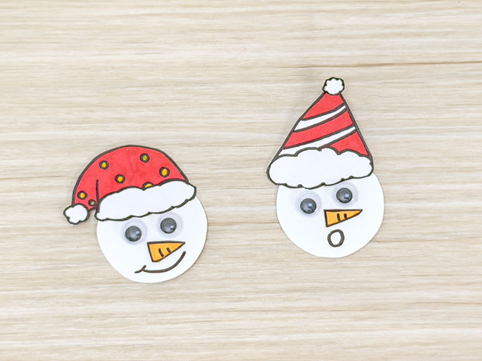 Two crafted snowman faces are displayed on a light wood-grained background. Each face is a white circle with googly eyes, an orange carrot nose, and a Santa hat. The snowman on the left wears a red Santa hat with yellow dots, and has a slight smile. The snowman on the right wears a red and white striped Santa hat and has a surprised expression with an open o shaped mouth. These appear to be handmade Christmas decorations.