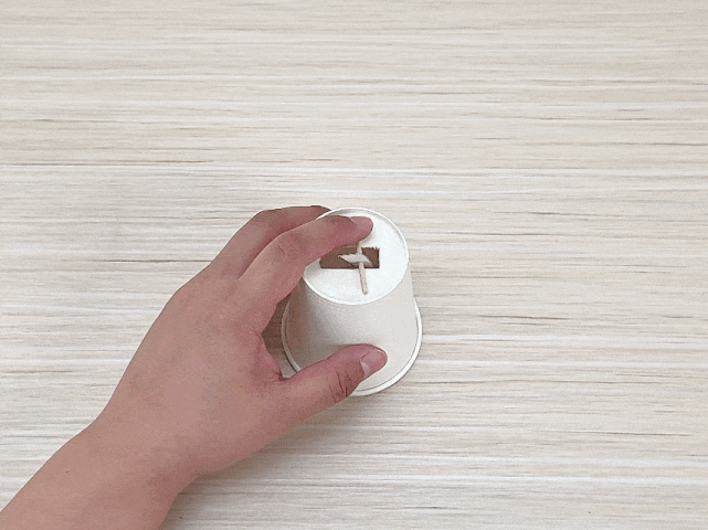 A hand interacts with a small, white paper cup placed upside down on a light wooden surface. A rectangular hole has been cut into the base of the cup, and a toothpick is inserted through it. The hand gently presses on the toothpick, seemingly testing a mechanism or construction involving the cup.