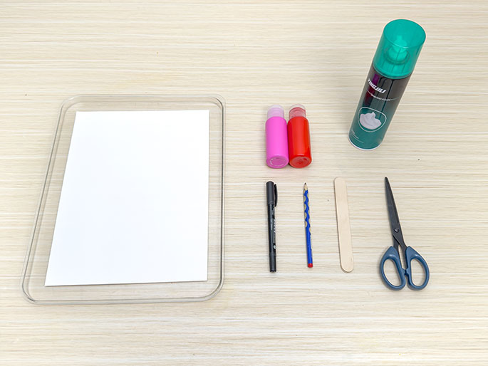 An overhead flatlay image displays art supplies arranged on a light wooden table. A clear plastic tray holds a blank white sheet of paper. Next to the tray are two small bottles of paint, one pink and one red, a can of shaving cream, a black marker, a blue pencil, a wooden craft stick, and a pair of scissors. These materials suggest preparation for a craft activity involving marbling or painting techniques.