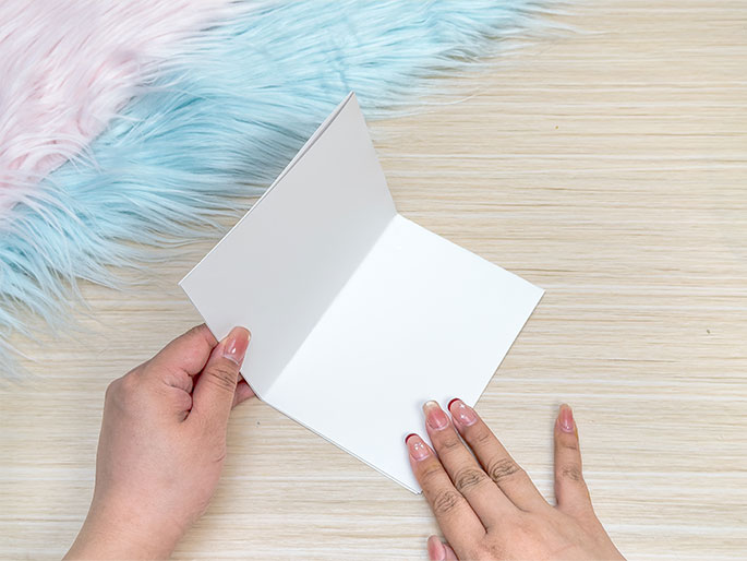 Two hands are holding open a blank white folded card, presumably to write in it. The card is positioned on a light wooden surface. A soft, fluffy material in shades of pink and teal is partially visible in the top left corner. The image suggests a stationery or card-making context.
