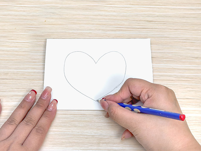 On a light woodgrain table, a hand holding a blue pencil is drawing a heart shape on a piece of white paper. Another hand is partially visible on the left side of the image. The focus is on the act of drawing the heart.