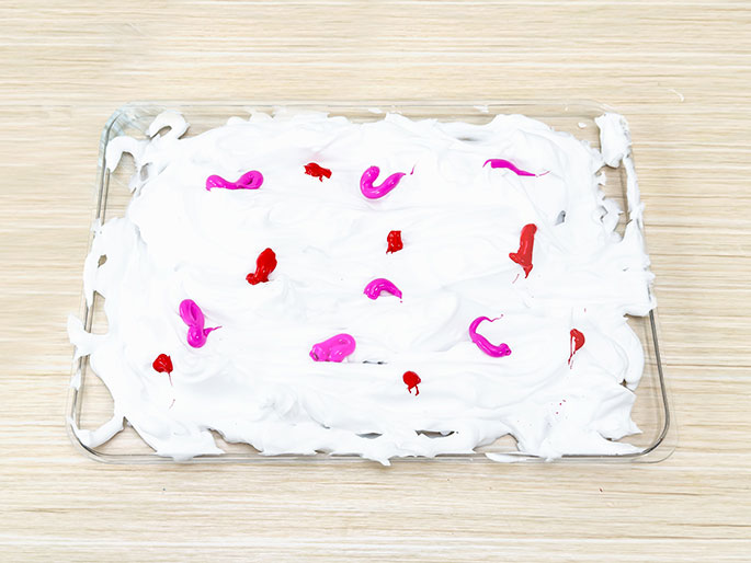 A clear tray containing a smooth layer of white shaving cream sits on a light wooden surface. Drops of pink and red paint have been added to the shaving cream, creating a pattern of color against the white background. This appears to be the setup for a marbling or painting activity.