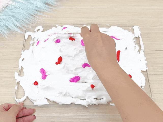A hand uses a craft stick to swirl pink and red paint drops into a layer of white shaving cream spread within a clear tray. The tray rests on a light wooden surface. A second hand is partially visible in the bottom left corner, holding the tray steady. This process appears to be creating a marbled effect in the shaving cream. A fluffy, light-colored material is visible in the top left corner.