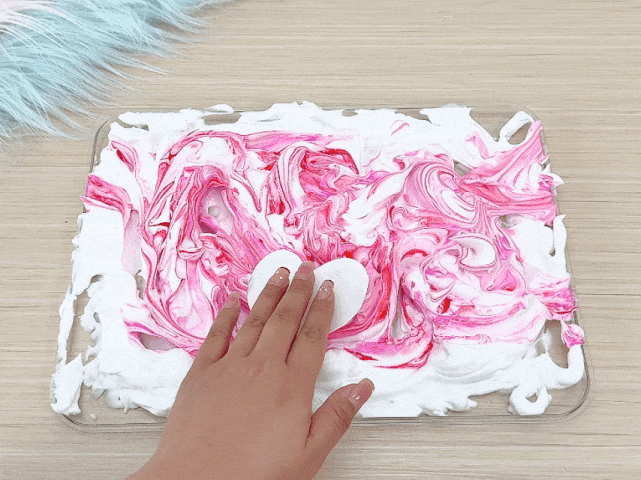 A hand presses a white heart-shaped cutout onto a layer of shaving cream marbled with pink and red paint. The shaving cream sits in a clear tray on a light wooden table. Some unmarbled white shaving cream is visible around the edges of the tray. A fluffy, light-colored material is partially visible in the top left corner. This image appears to depict a stage in a marbling craft project.