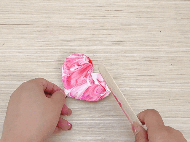 Two hands are visible on a light woodgrain surface. One hand holds a white heart shape, now marbled with pink and red paint, presumably transferred from the shaving cream marbling process seen in previous images. The other hand uses a wooden craft stick to scrape excess shaving cream from the marbled heart.