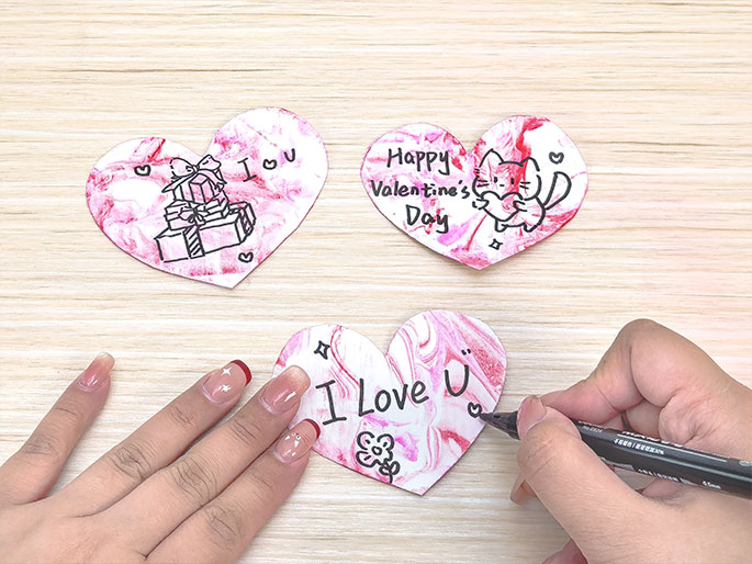 Three heart-shaped cards rest on a light woodgrain surface. Each heart features a marbled pink and white design, created with paint and shaving cream. The first heart has a drawing of stacked gifts and the words I ♡ U. The second heart reads Happy Valentines Day and features a drawing of a cat holding a heart. A hand is seen writing on the third heart, which has a small flower drawing and the partially completed phrase I Love U.