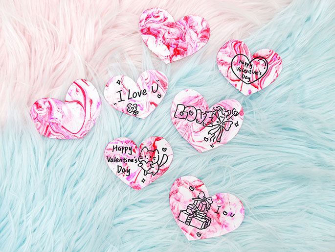 Six hand-decorated Valentine's Day hearts lie on a fluffy, light blue and pink background. Each heart is white with a marbled pink design and features simple black ink drawings and messages. Two of the hearts say Happy Valentines Day, one with a drawing of a cat holding a heart, the other with small stars. Two hearts have drawings of stacked presents and the message I ♡ U. One heart has the message I Love U with a small flower, and the final heart has the word LOVE with a drawing of a ribbon-tied bouquet and small stars.
