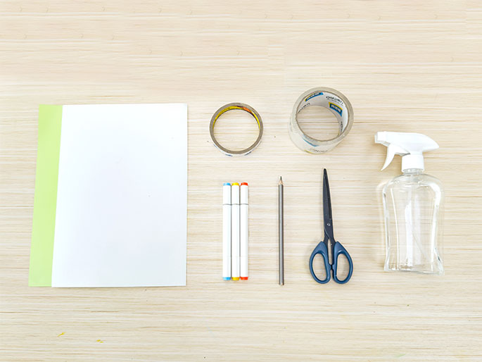 An assortment of craft supplies lies arranged on a light wooden table. These supplies include a piece of white paper with a light green strip attached to the side, two rolls of clear tape (one started and one new), three fine-tipped markers (blue, white, and orange), a pencil, a pair of scissors, and a clear spray bottle. These items appear to be the materials needed for a craft project.