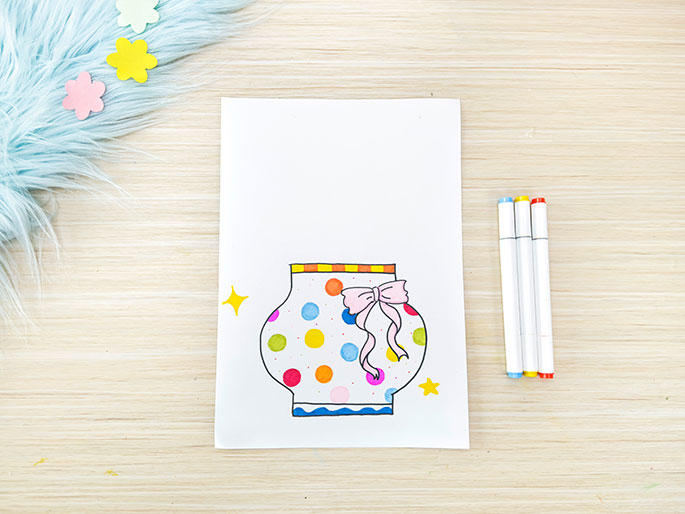 A piece of white paper on a light wooden table displays a drawing of a rounded vase or jar. The vase is decorated with colorful polka dots and a pink ribbon tied around its neck. A striped orange and yellow band adorns the top of the vase, and small, sparkly yellow stars are drawn on either side. Three white markers with orange, blue, and yellow caps lie next to the paper. A piece of fluffy light blue material with small pastel flower shapes is visible in the top left corner. This image appears to depict a stage in a children's craft project.