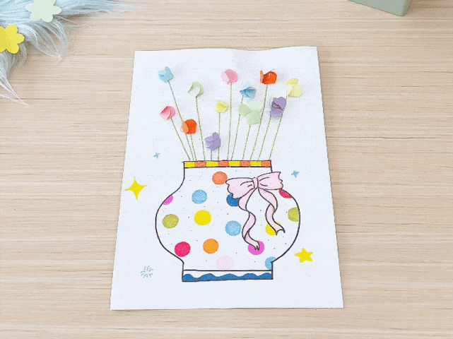 A finished children's craft project rests on a light wooden table. The project, created on a sheet of white paper, depicts a vase filled with colorful flowers. The vase is adorned with brightly colored polka dots and a pink ribbon tied around its neck. Small, folded paper flowers in various pastel shades are attached to thin green stems that rise from the vase's opening. Small yellow stars are drawn around the vase, adding to the decorative touch. A piece of fluffy light blue material with pastel flower shapes is partially visible in the top left corner. The image captures the charm and simplicity of a child's artwork.