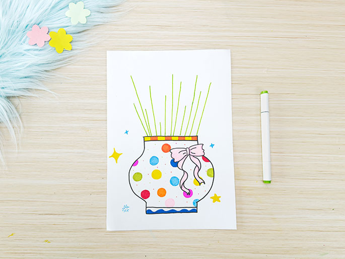 A drawing of a vase with flowers sits on a piece of white paper placed on a light wood-grain surface. The vase is decorated with colorful polka dots, a pink ribbon tied around the neck, and an orange and yellow striped band at the top. Green lines, representing flower stems, extend upwards from the vase's opening. Small, sparkly yellow stars are drawn on either side. A green marker lies beside the paper, suggesting it was used to draw the stems. A fluffy, light blue material with small pastel flower shapes is partially visible in the top left corner. This image appears to be of a children's art project in progress.