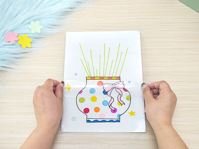 Two hands are applying a sheet of clear adhesive plastic to a piece of white paper. The paper, resting on a light wood-grain surface, features a drawing of a vase filled with green stems. The vase is decorated with colorful polka dots, a pink ribbon, and an orange and yellow striped band. Small yellow stars are drawn on either side. A piece of light blue fluffy material with pastel flower shapes is partially visible in the top left corner. The image seems to depict a stage in a children's craft project, where the drawing is being covered for protection or to create a sticker-like effect.