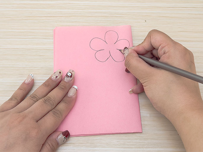 On a light wooden table, two hands are shown interacting with a folded piece of light pink paper. One hand holds the paper steady while the other hand draws a simple flower shape with a pencil onto the paper. The hand drawing has decorated fingernails. The image suggests a craft activity, likely card-making or a similar paper project.
