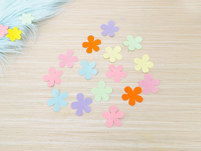 Several small, simple flower shapes cut from pastel-colored paper are scattered on a light wooden surface. The flowers are in various shades of pink, orange, yellow, green, and purple. A piece of fluffy light blue material is partially visible in the top left corner, also adorned with a few small paper flowers. The image evokes a sense of spring or crafting.