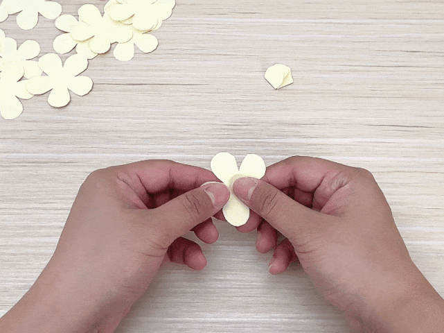 Two hands are manipulating a small, pale yellow paper flower shape against a light wood background. Several more of these paper flower cutouts are visible at the top of the frame. The hands are pinching the base of the flower, seemingly in the process of folding or shaping it. A smaller, folded piece of the same paper sits to the upper right, suggesting a step in a craft project involving paper flowers.