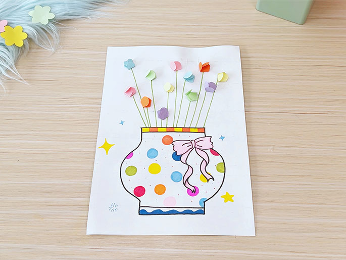 A piece of white paper on a light wood-grain table displays a children's craft project. Drawn on the paper is a vase decorated with colorful polka dots, a pink ribbon, and an orange and yellow striped rim. Small yellow stars are drawn on either side. Sticking out of the vase are thin green stems with small, folded paper flowers in various pastel colors attached. A piece of light blue fluffy material with pastel flower shapes is partially visible in the top left corner. The image depicts a completed flower-in-a-vase craft.