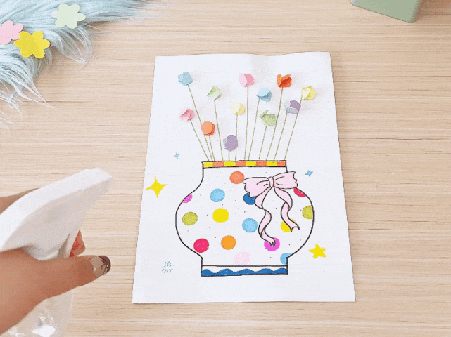 A hand holding a spray bottle is positioned to the left of a children's craft project on a light wooden table. The project, displayed on white paper, features a drawing of a vase decorated with colorful polka dots and a pink ribbon. Small, folded paper flowers in various pastel colors are attached to thin green stems growing from the vase. Small yellow stars are drawn around the vase. A piece of light blue fluffy material and more pastel flower shapes are partially visible in the top left corner. It appears the spray bottle is about to be used on the craft, possibly containing adhesive or a sealant.