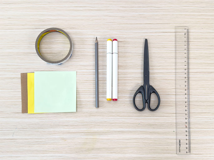 An assortment of craft supplies is arranged on a light woodgrain table or desk. From left to right: a roll of clear tape, three small, stacked squares of paper (brown, yellow, and light green), a gray pencil, two white markers with red and yellow caps, a pair of black scissors, and a clear plastic ruler. The image shows the materials needed for a craft project.