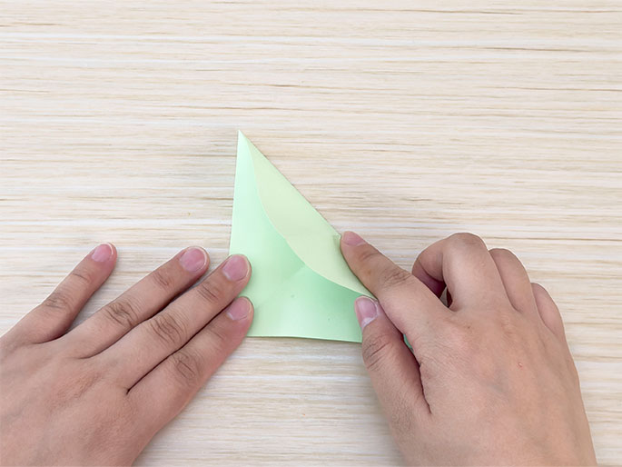 Two hands fold a piece of light green paper into a triangle shape on a light woodgrain surface. The fingers hold the paper as it's being creased. The image demonstrates a step in a paper folding or origami process.