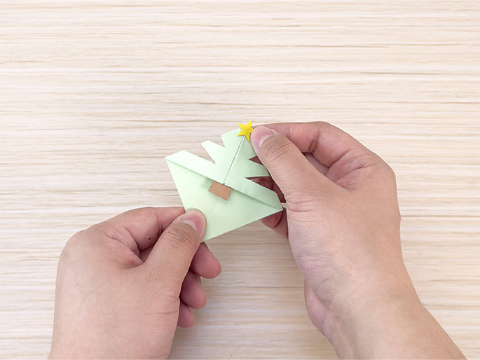 Two hands hold a small, light green origami Christmas tree against a light woodgrain background. The tree is decorated with a small, yellow star at the top and a small, brown square attached to the trunk area. The hands are carefully positioning or adjusting the tree.