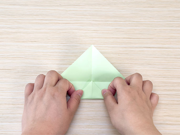 Two hands fold a light green piece of paper into a smaller triangle shape on a light woodgrain surface. The paper has been folded in half to create a triangle, and now the outer corners are being folded in to meet at the center crease, creating a narrower, kite-like shape. The image shows a step in a paper folding or origami process.