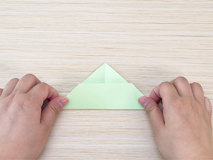 Two hands hold a light green piece of paper folded into a triangular shape with a rectangular base on a light woodgrain surface. The paper has been folded several times to create this shape, which resembles a simple house or a tree. The image shows a step in a paper folding or origami process.
