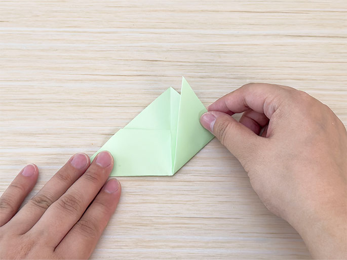 Two hands are folding a piece of light green paper on a light woodgrain surface. The paper is already folded into a triangular shape, and one of the outer flaps is being folded inwards towards the center crease. This action is creating a smaller, layered triangular form. The image shows a step in a paper folding or origami process.