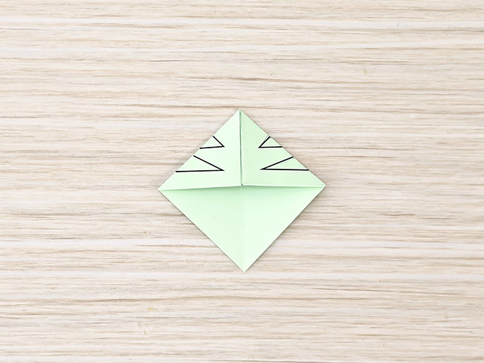 A light green piece of folded paper sits on a light woodgrain surface. The paper is folded into a diamond shape, and the upper flaps have small, symmetrical black lines drawn on them, creating a decorative pattern. The image showcases a stage in an origami or paper folding activity.