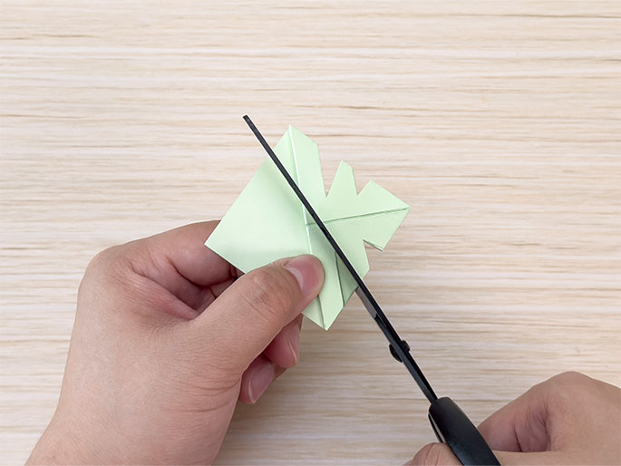 A pair of scissors with black handles cuts into a folded piece of light green paper held by two hands against a light woodgrain background. The paper is folded into a complex shape with several triangular sections, and the scissors are making a cut into one of these sections. This image illustrates a step in a crafting or origami project.