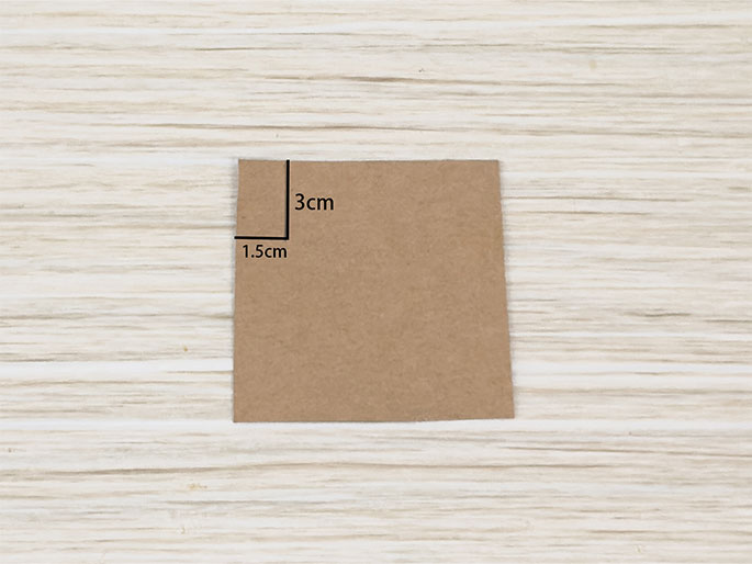 A small square of brown paper rests on a light woodgrain surface. The square measures 3cm x 1.5cm, as indicated by black lines and text labels along two of its sides. The image likely depicts a piece of paper being prepared for a craft or origami project.