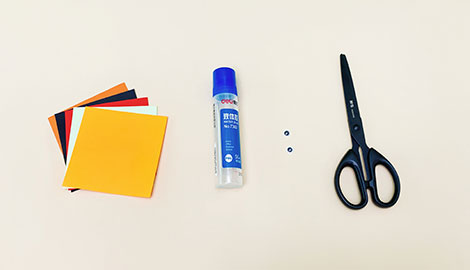 The image shows the materials needed to make an origami bat bookmark. Laid out on a pale beige surface are a stack of coloured square papers (including orange, yellow, black, red and white), a small bottle of glue, two small, dark-coloured googly eyes, and a pair of black scissors. The arrangement is neat and clearly shows all the necessary components for the craft project. The colours are bright and cheerful, suggesting a fun and creative activity.