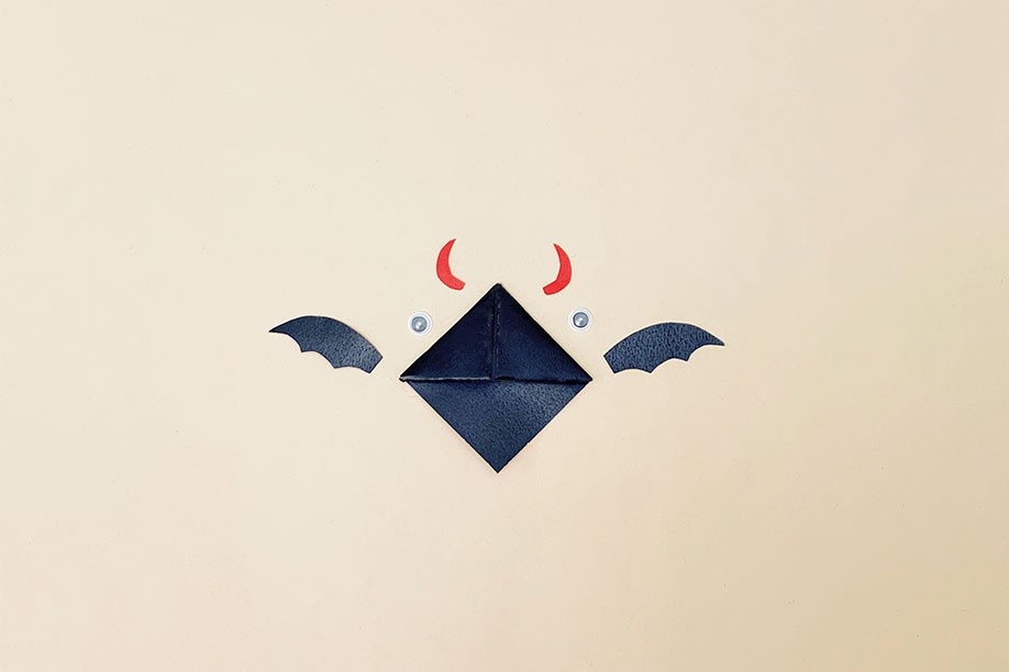 A playful, Halloween-themed craft is depicted: a folded black square of card forms the body of a bat, with added black paper wings and small red horns at the top. Two white googly eyes are positioned centrally, completing the whimsical design. The components are arranged on a pale beige background, creating a simple yet effective visual representation of the finished bookmark. The overall impression is one of fun, creativity, and a simple craft project perfect for Halloween.